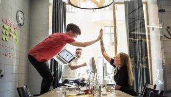Et team av to unge menn og en ung kvinne ler og gir hverandre high five rundt et stort bord på et kontor.