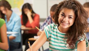 Ung kvinnelig elev sitter smilende og skriver i et klasserom.